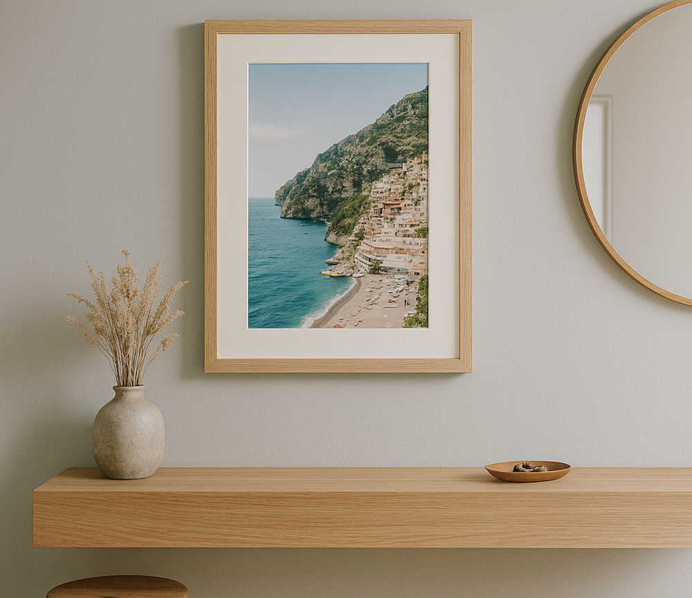 A2 natural oak picture frame with matboard displaying a Mediterranean coastal print, mounted above a floating timber console in a modern hallway with light grey wall, round mirror, and natural decor in soft daylight.