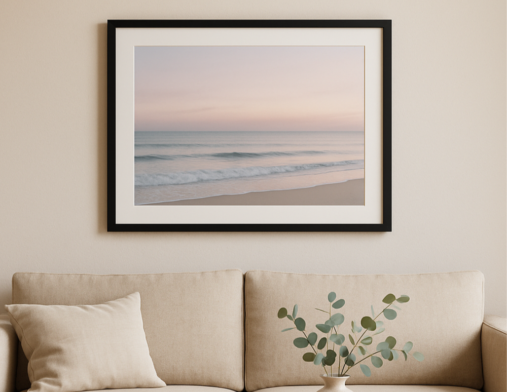 A black A2 frame with white matboard displaying a pastel coastal photo, mounted above a beige sofa in a minimalist living room with eucalyptus decor and soft natural light.
