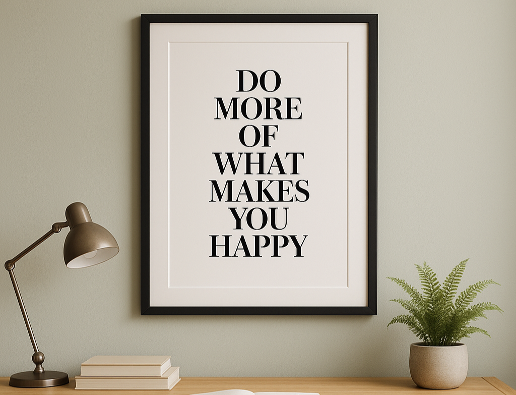 A black A2 frame with white matboard showcasing a bold typographic quote, hanging above a light wood desk with books, a notepad, and a fern plant against a sage green wall.