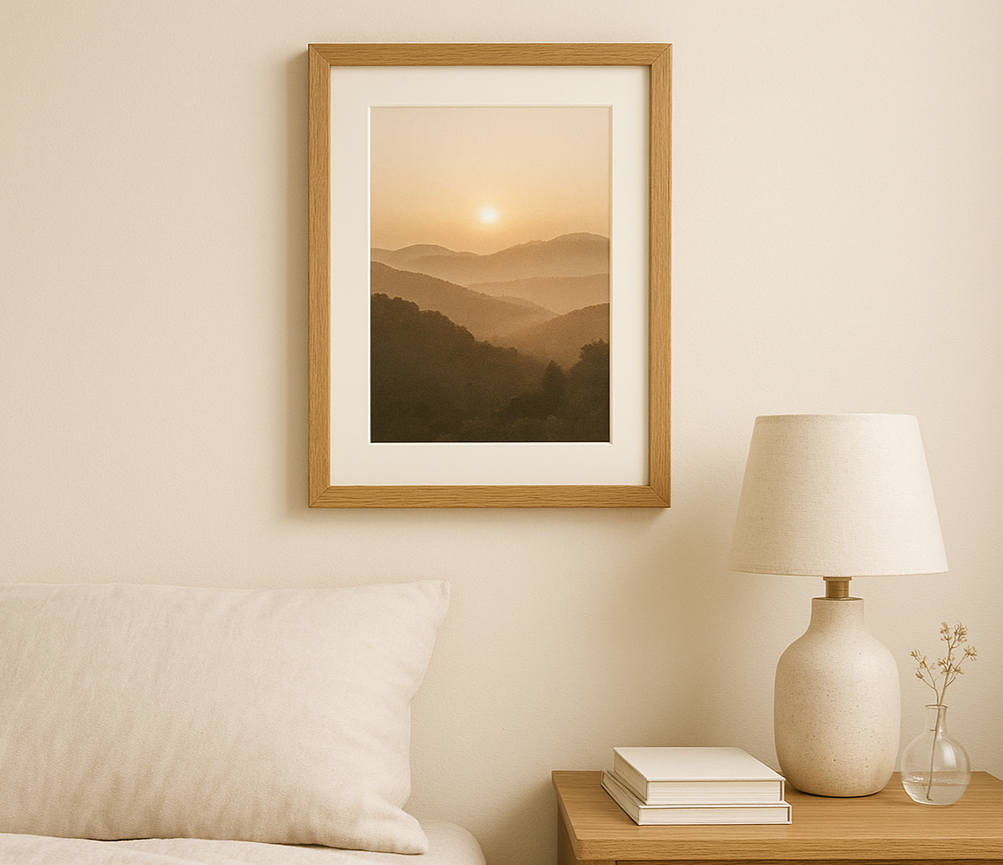 A3 premium oak picture frame with matboard featuring a golden sunrise over hills, mounted above an oak bedside table in a minimalist bedroom with linen bedding and soft lighting.